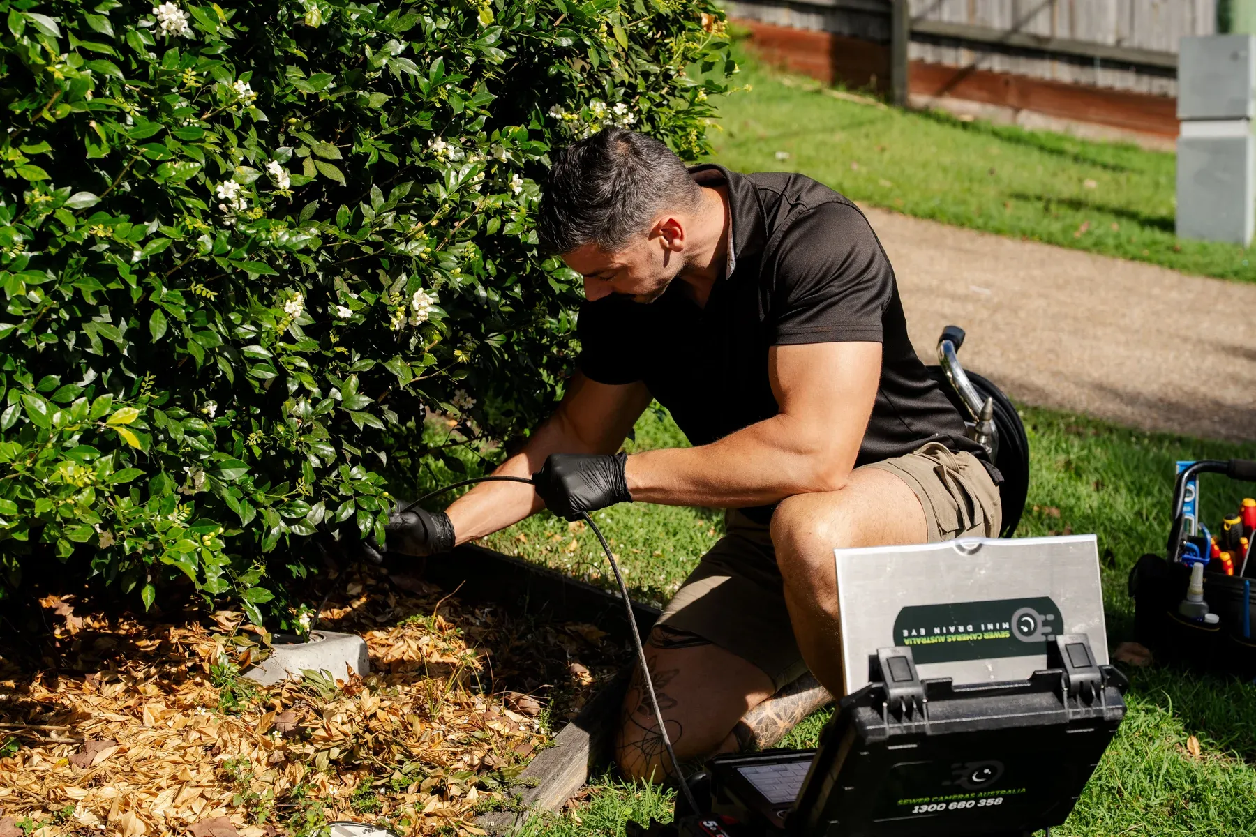 Plumber feeding a drain camera cable into an access point beside a garden